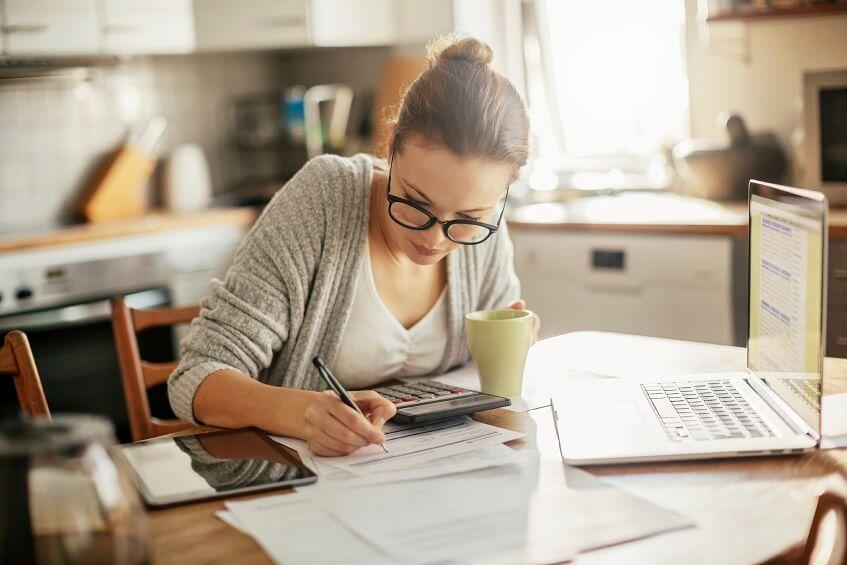 A woman in glasses busily using a laptop and pen to complete work.