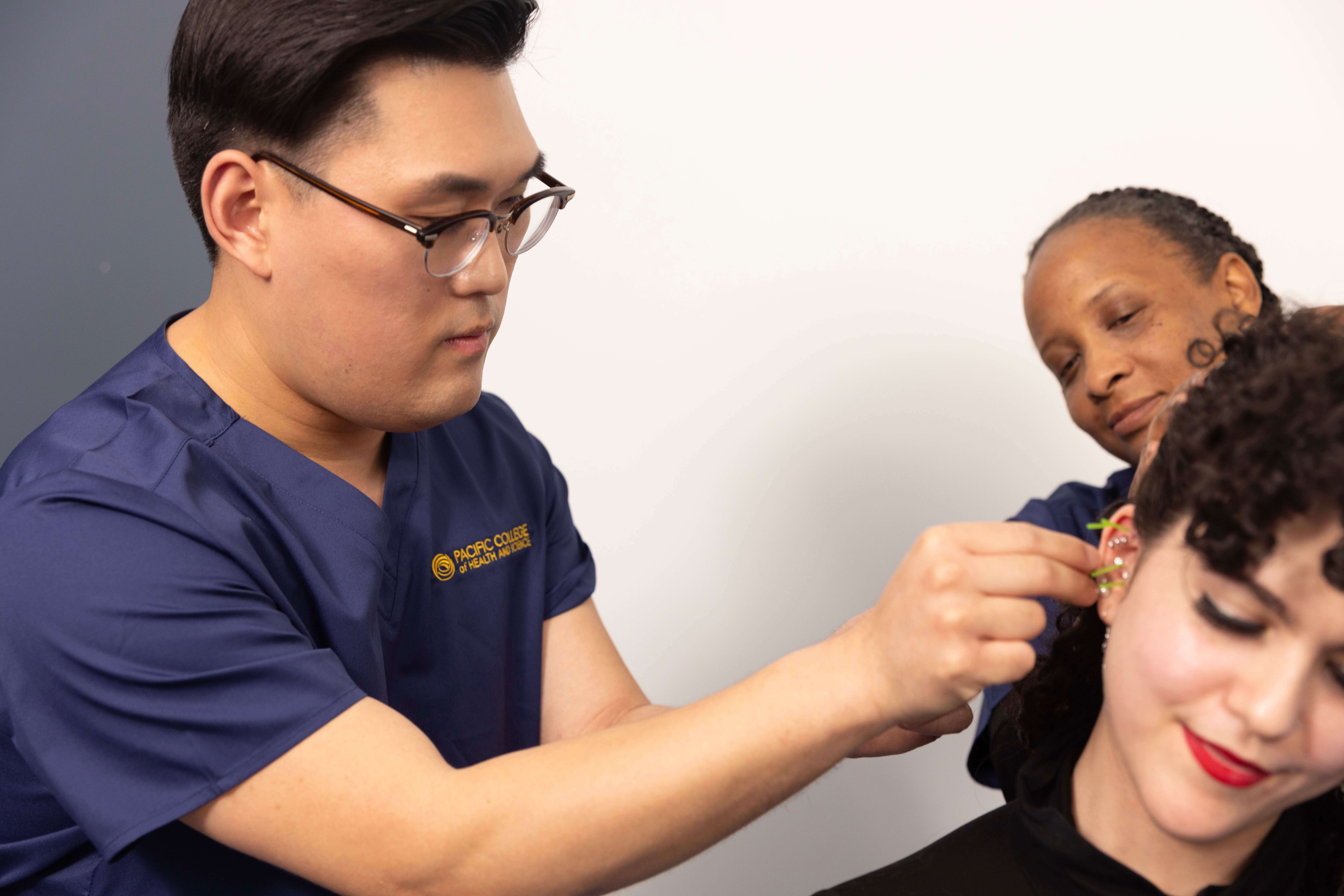 Acupuncture student performing ear acupuncture treatment while instructor observes during clinical training